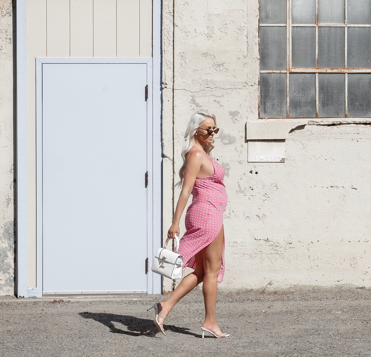 Fashion blogger walking, wearing sunglasses, a pink poka dot midi dress, and white kitten heels